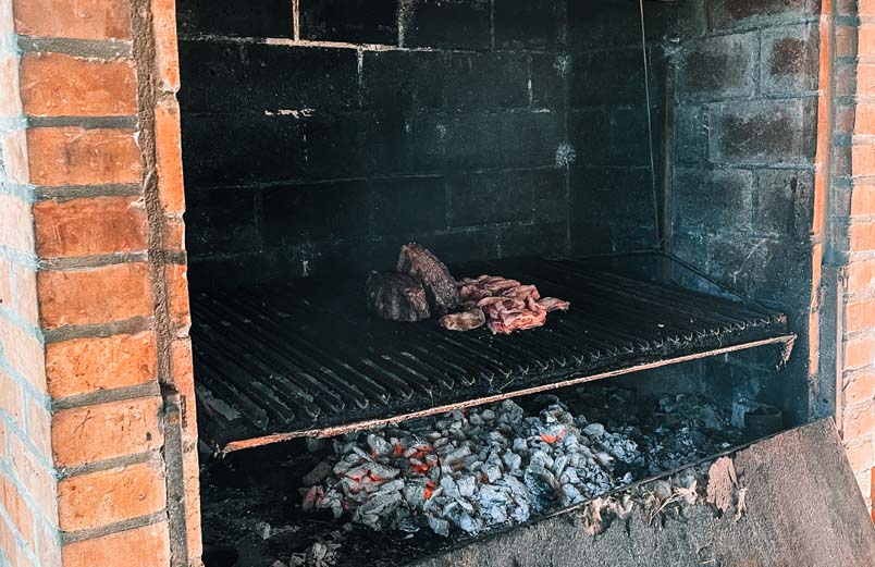 Meat on a grate over smoldering embers in a large outdoor fireplace