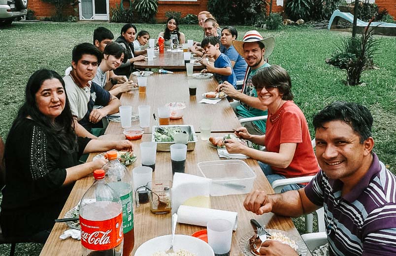 Thirteen people eating around three tables outside on a green lawn