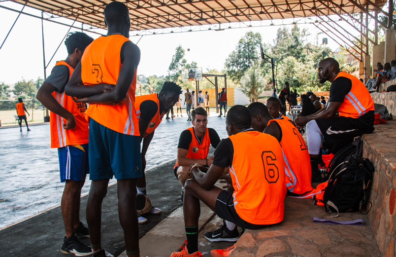 A group of basketball players in orange jerseys talk in a circle on the sidelines with a game going on in the background.