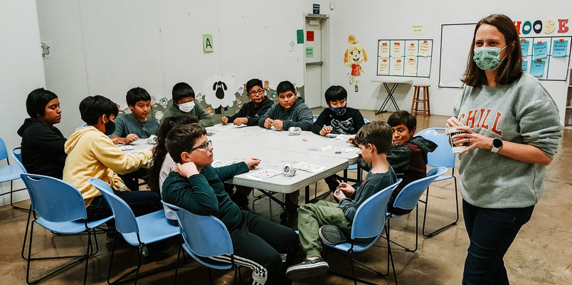 Ashley with children at The Center A woman in a mask stands next to a table where children are seated with papers in front of them.