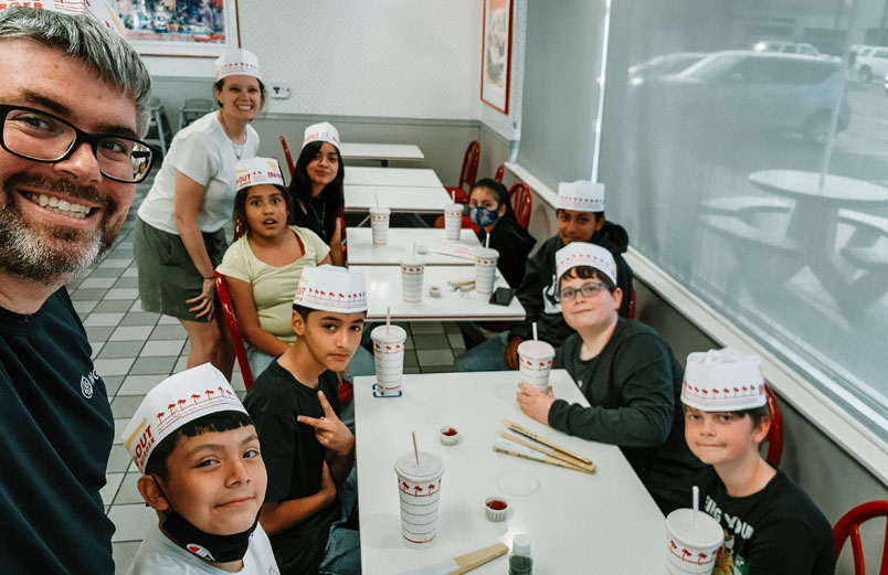 A husband and wife with a group of children inside a restaurant, all wearing paper hats on their heads.