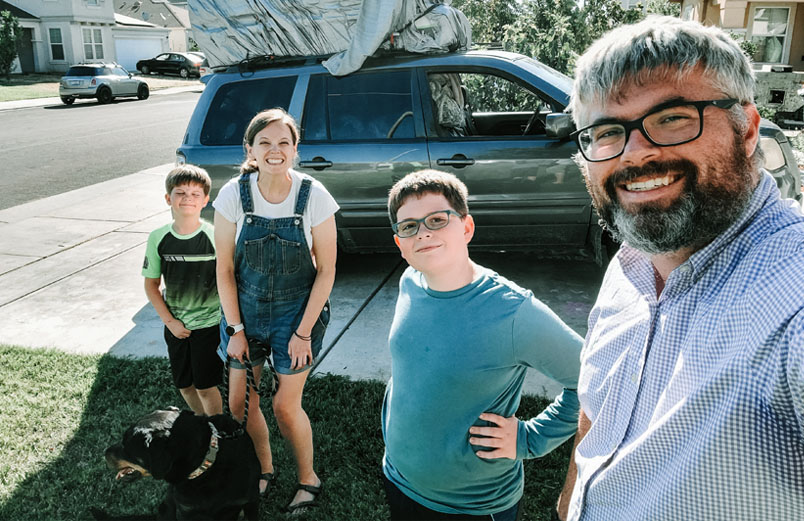 A husband and wife pose with their two boys and their dog in front of their van in the driveway.