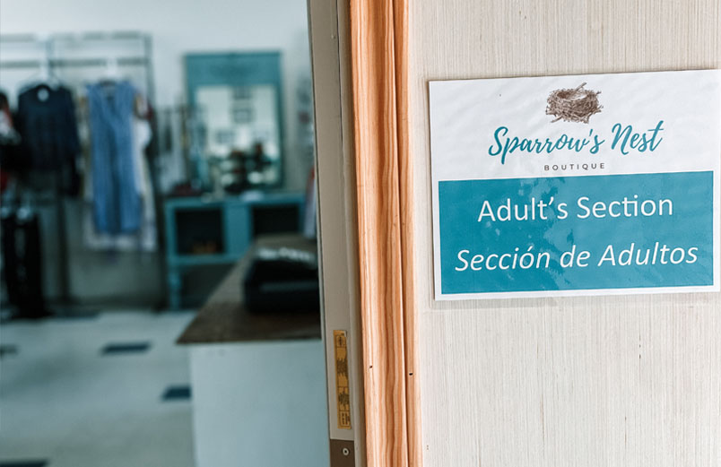 Clothes hang on racks in the background, with a sign reading “Sparrow’s Nest Boutique, Adult’s Section, Sección de Adultos” in the foreground.