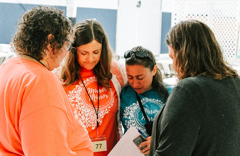 Four women stand in a circle with their heads bowed and their eyes closed, praying.