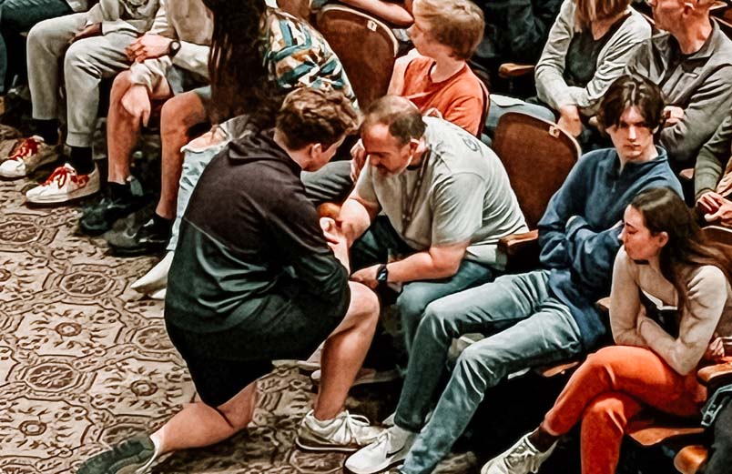 A young man kneels with his head bowed, facing his his father in a crowded auditorium