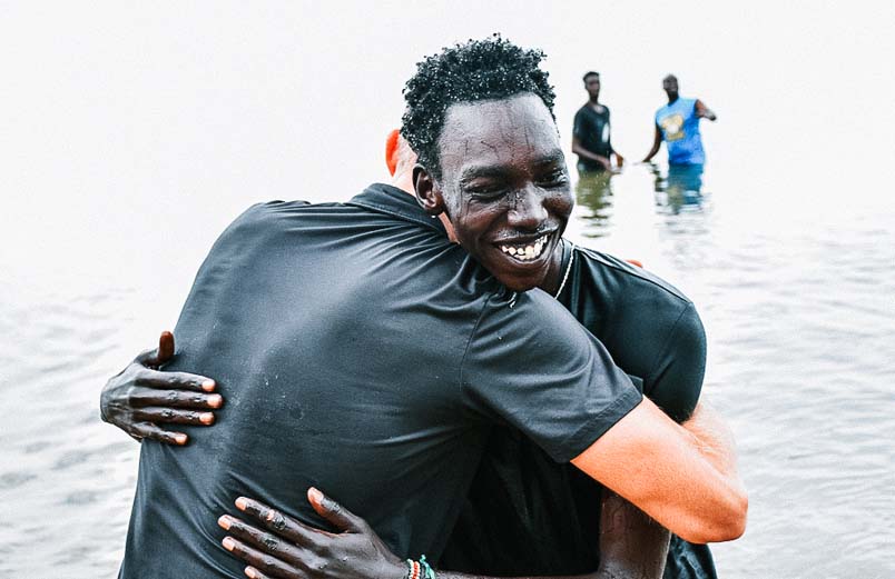 A man wet from his baptism on the shore of the lake hugging a friend.