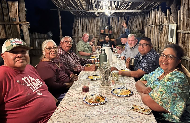 A group of people sit at a table under a ramada, eating a meal.