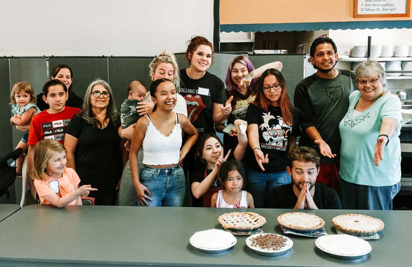 A group of smiling people standing behind a table displaying six homemade pies of differing types.