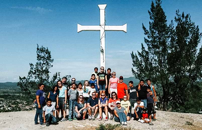 A group of smiling people in front of a large white cross standing on a hill overlooking a village..