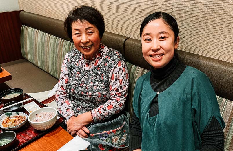 Two women, smiling for the camera, sitting side by side in a booth with their meals in front of them.