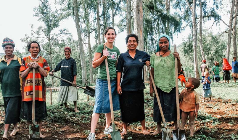 Veronica and five women stopping work and posing with their shovels along with children and others in the background.